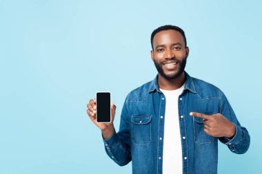 Neşeli Afrikalı Amerikalı adam mavi ekranda boş ekranlı akıllı telefonu işaret ediyor.