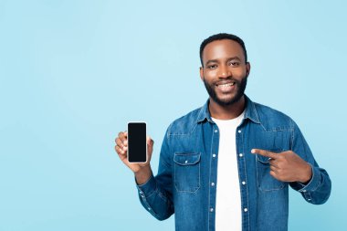 cheerful african american man pointing at smartphone with blank screen isolated on blue clipart