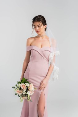 Pembe elbiseli ve peçeli genç gelin gri renkte izole edilmiş düğün buketini tutuyor.