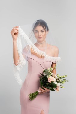 Duvağa dokunan şehvetli kadın, gri renkte izole edilmiş düğün buketini tutuyor.