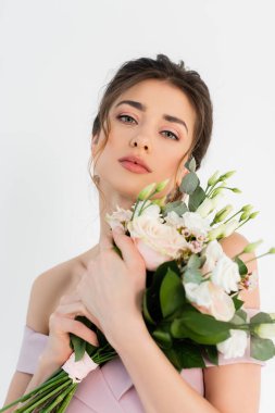 Genç gelin kameraya bakarken düğün buketini beyazın üzerinde soyutlanmış halde tutuyor.