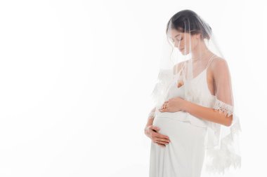 Gelinlik ve duvak takmış hamile bir kadın göbeğine dokunuyor.