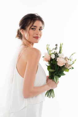 Genç gelin kameraya bakarken düğün buketini beyazın üzerinde soyutlanmış halde tutuyor.