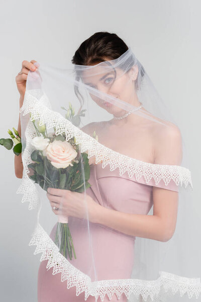 flirty woman covering face with veil while standing with wedding bouquet isolated on grey