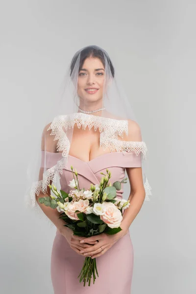 pretty woman in pink dress and veil holding wedding bouquet isolated on grey