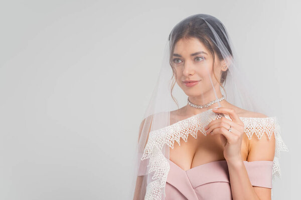 smiling bride touching veil and looking away isolated on grey