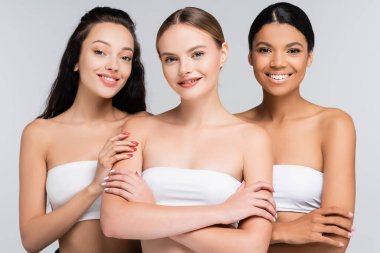happy multiethnic women with bare shoulders in tops isolated on grey clipart
