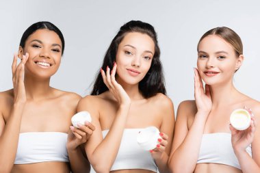 Çıplak omuzlu çok ırklı kadınlar ellerinde kaplar tutarken gülümsüyor ve gri renge kozmetik krem sürüyorlar.