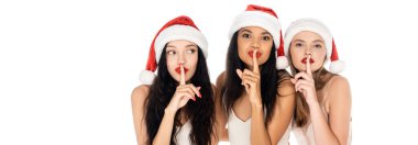 Beyaz pankartta izole edilmiş sus işareti gösteren Noel Baba şapkalı genç ırklar arası kadınlar.