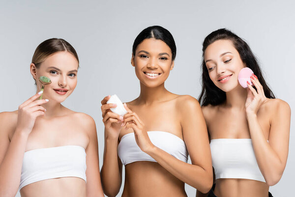 счастливые многоэтнические женщины, держащие контейнер с косметическим кремом, нефритовый роллер и силиконовый очиститель изолированы на сером