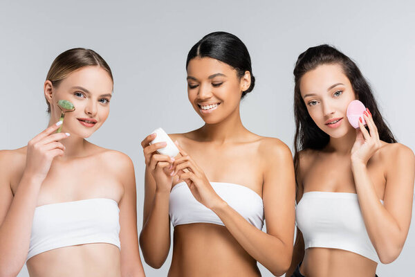 cheerful multiethnic women smiling while holding container with cosmetic cream, jade roller and silicone cleanser isolated on grey
