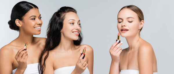 multiethnic women with bare shoulders smiling while holding lipsticks isolated on grey, banner