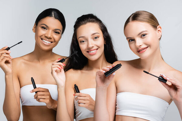 multiethnic women with bare shoulders smiling while holding mascara tubes isolated on grey