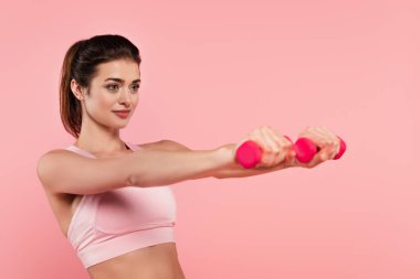 Formda sporcu kadın, dambıllarla egzersiz yapıyor. Önü bulanık. Pembe renkte. 