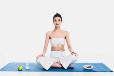 Yoga yapan kadın elmanın yanında oturuyor, beyaz arka planda bir bardak su ve mısır gevreği. 