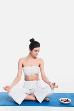 Genç bir kadın mısır gevreği kasesinin yanında meditasyon yapıyor. Beyaz arka planda spor minderinin üstünde böğürtlen var. 