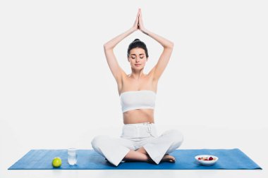 Esmer kadın mısır gevreğinin yanında meditasyon yapıyor, elma ve su beyaz arka planda fitness minderi üzerinde 