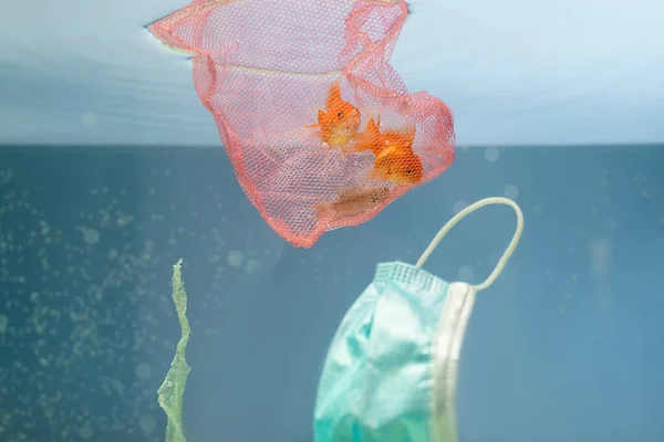 stock image goldfishes in net, used medical mask and cellophane rubbish in water, ecology concept