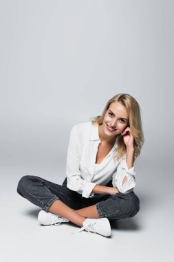 happy blonde woman smiling at camera while sitting with crossed legs on grey