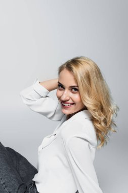 happy blonde woman holding hand near head while sitting isolated on grey