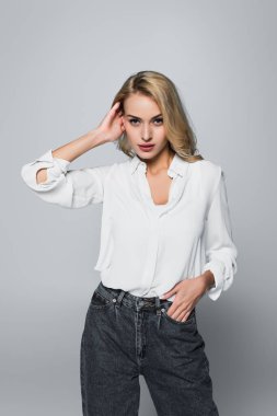 pretty, blonde woman in white shirt touching hair while posing with hand in pocket on grey