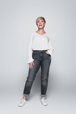 full length view of mature woman in white blouse and jeans standing with hand in pocket on grey