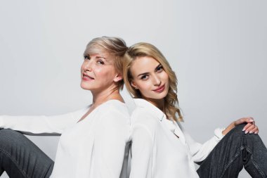 trendy mother and daughter smiling at camera while sitting isolated on grey