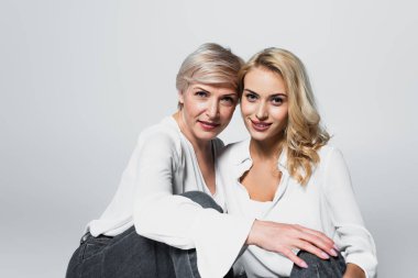 stylish mother and daughter smiling at camera while sitting isolated on grey