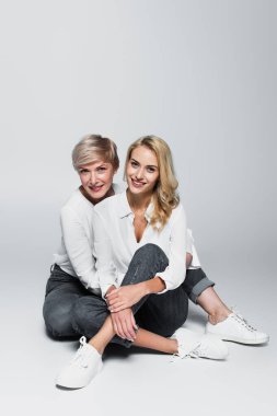 full length view of trendy mother and daughter smiling at camera while sitting on grey