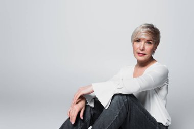 mature woman in white blouse and jeans looking at camera while sitting isolated on grey