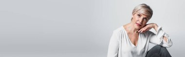 mature woman in white blouse posing with hand near face isolated on grey, banner