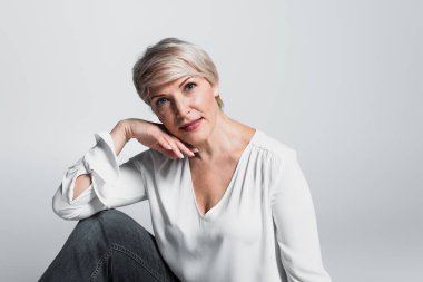 stylish, mature woman looking at camera while holding hand near face isolated on grey