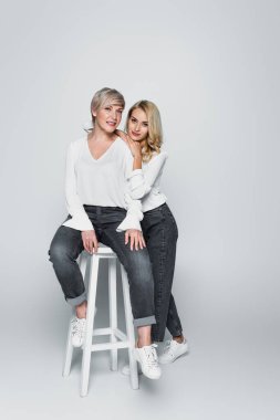 full length view of young stylish woman leaning on smiling mother sitting on high chair on grey