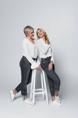 full length view of trendy mother and daughter looking at each other near high chair on grey