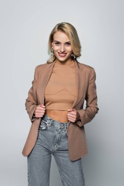 cheerful, trendy woman smiling at camera while posing on grey