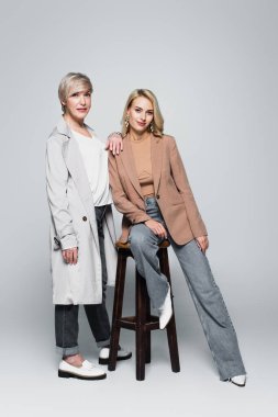 fashionable woman near stylish daughter sitting on high chair on grey