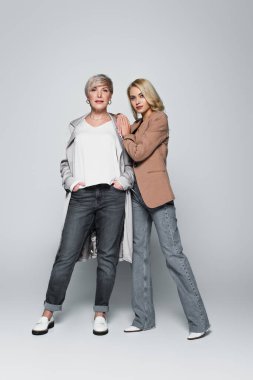 young woman leaning on shoulder of stylish mother posing with hands in pockets on grey