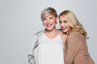 cheerful young woman leaning on shoulder of happy mother isolated on grey