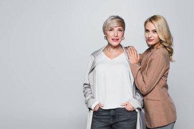 middle aged, trendy woman posing with hands in pockets near daughter isolated on grey