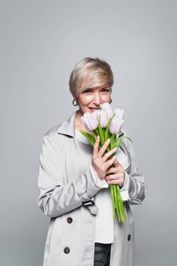 happy woman in stylish trench coat smelling fresh tulips isolated on grey