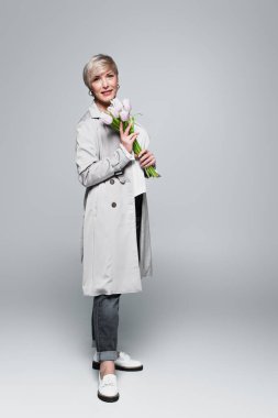 full length view of fashionable, mature woman holding tulips on grey