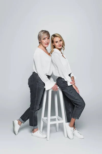 full length view of mother and daughter in jeans and white blouses posing near high chair on grey