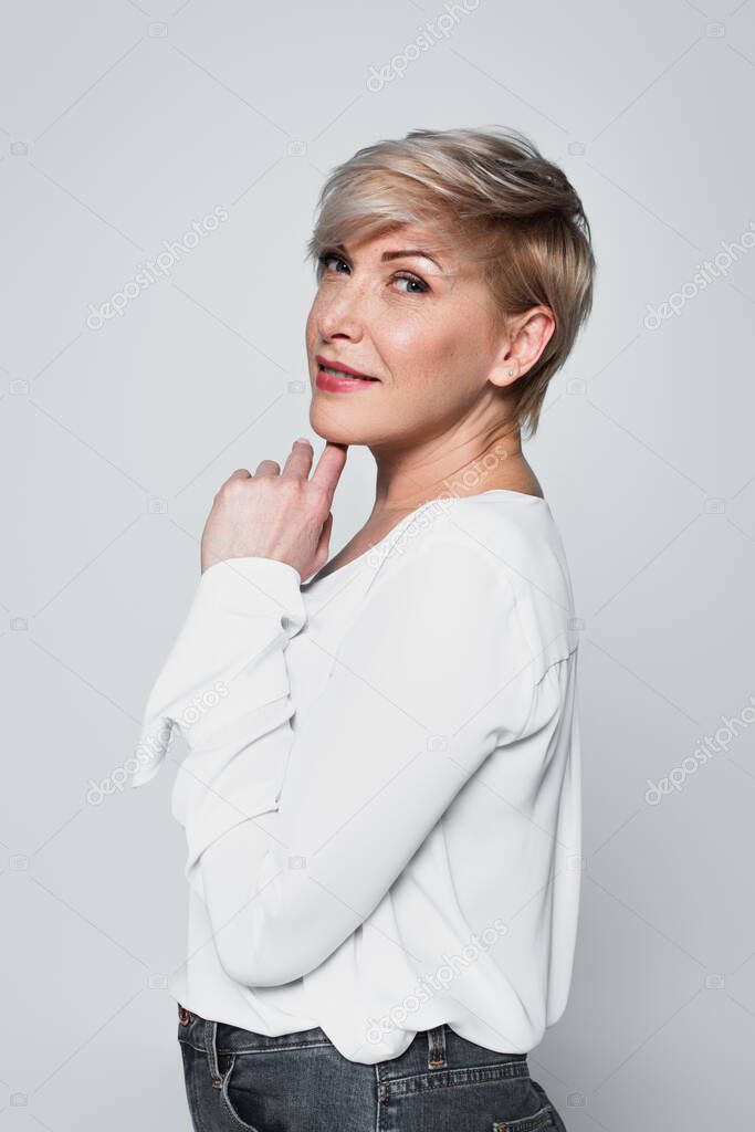 ash-blonde woman in white blouse posing with hand near chin isolated on grey
