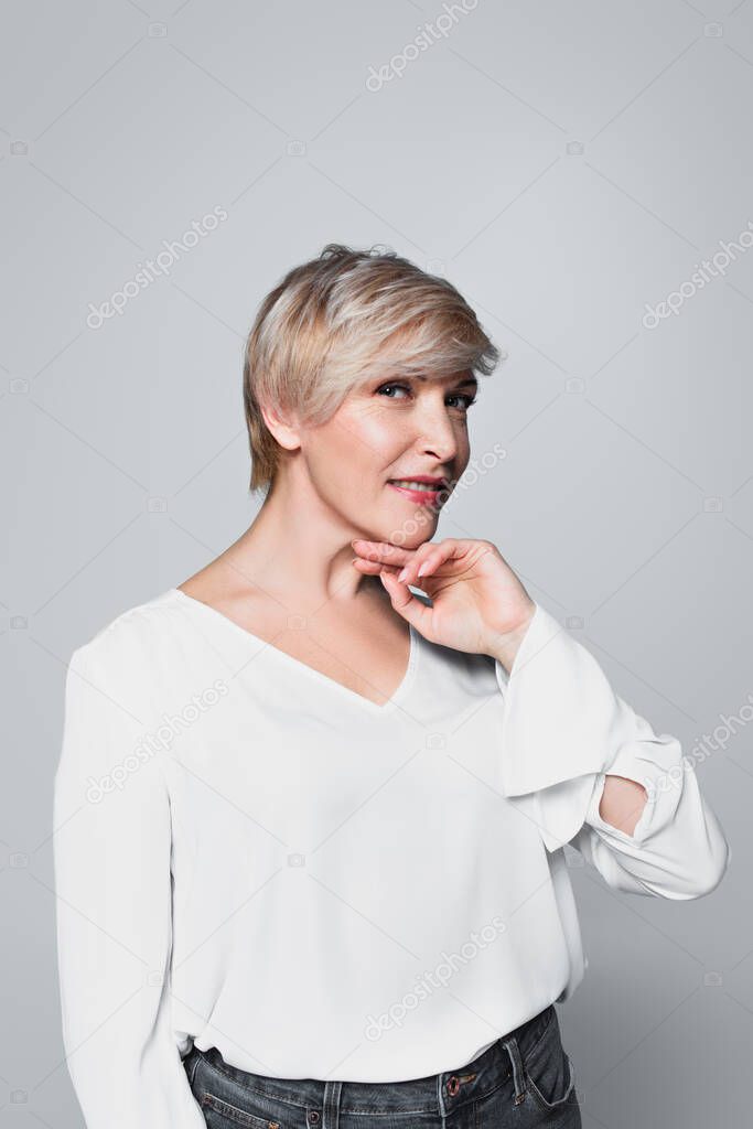 ash-blonde woman in white blouse posing with hand near chin isolated on grey
