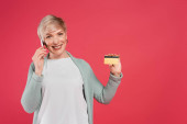  pleased, mature woman showing credit card while talking on smartphone isolated on pink