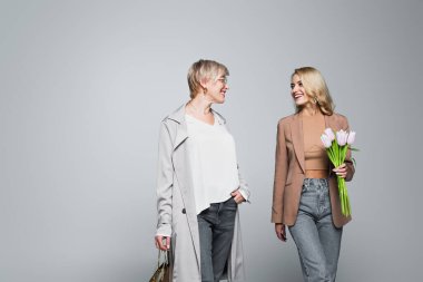 happy mother and daughter with shopping bag and tulips looking at each other on grey