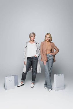 mother and daughter in trendy clothes posing near shopping bags on grey