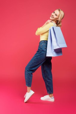 full length view of happy, stylish woman posing with shopping bags on pink