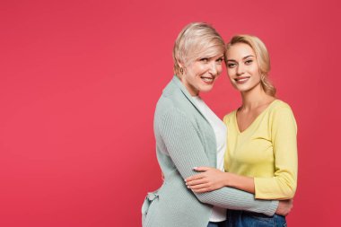 cheerful mother and daughter embracing while looking at camera isolated on pink