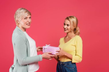 happy middle aged woman taking gift from adult daughter while looking at camera isolated on pink